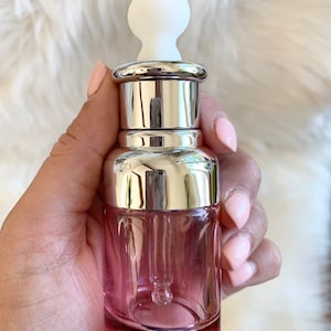 Puede incluir: Una botella de vidrio transparente con un tinte rosado y una tapa plateada. La botella tiene un gotero unido a la tapa. El gotero est&aacute; hecho de vidrio transparente y tiene una punta de goma blanca.