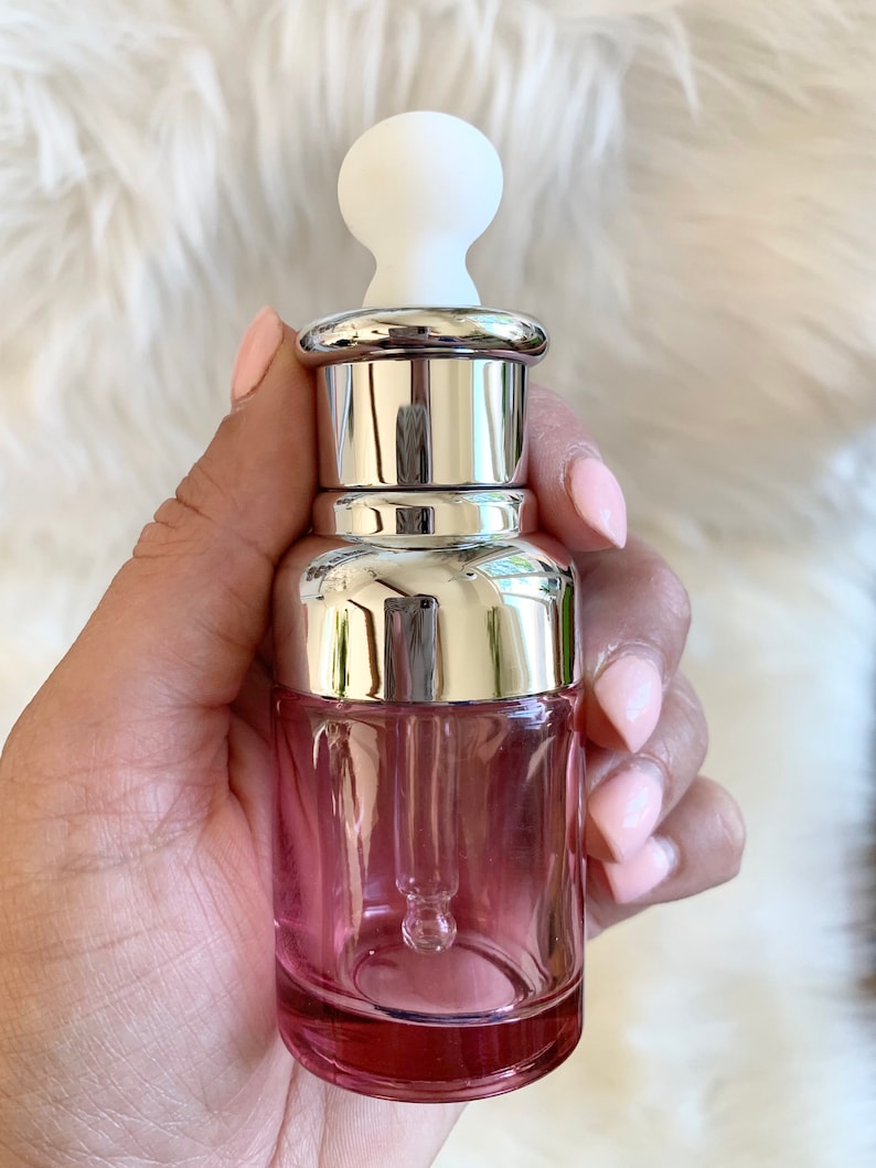Puede incluir: Una botella de vidrio transparente con un tinte rosado y una tapa plateada. La botella tiene un gotero unido a la tapa. El gotero est&aacute; hecho de vidrio transparente y tiene una punta de goma blanca.