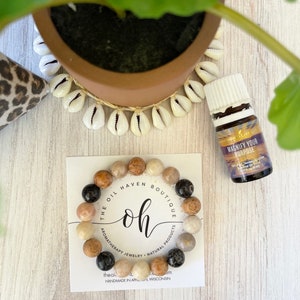Può includere: Un braccialetto di perline di pietra di vari colori, tra cui nero, beige e marrone, è appoggiato su un biglietto bianco. Il biglietto recita "THE OIL HAVEN BOUTIQUE" e "oh". Nella foto sono presenti anche una piccola bottiglia di olio essenziale e una pianta in vaso.