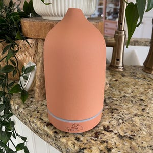Puede incluir: Un difusor de aceites esenciales de color terracota con una parte superior redondeada y una base iluminada. El difusor tiene el texto "Oil the Haven" impreso. El difusor est&aacute; sobre una encimera de granito.