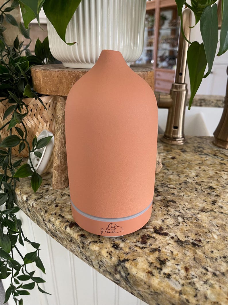Puede incluir: Un difusor de aceites esenciales de color terracota con una parte superior redondeada y una base iluminada. El difusor tiene el texto "Oil the Haven" impreso. El difusor est&aacute; sobre una encimera de granito.