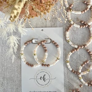 Pode incluir: Um par de brincos grandes de argola feitos com madeira natural e contas brancas. Os brincos são exibidos em um cartão branco com o texto "The Oil Haven Boutique" e "oh" em um círculo. Os brincos são feitos à mão em Appleton, Wisconsin.