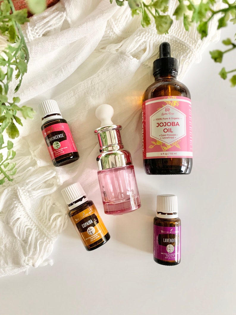 Puede incluir: Cinco botellas de aceite esencial con tapas y etiquetas blancas. Las botellas est&aacute;n sobre una superficie blanca con una tela de crochet blanca en el fondo. Las etiquetas dicen "Frankincense", "Copaiba", "Lavender" y "Jojoba Oil".