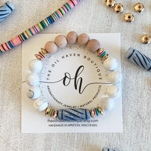 Puede incluir: Un brazalete de mármol blanco y gris con cuentas de madera, cuentas doradas y una cuenta rayada azul y blanca. El brazalete está sobre una tarjeta blanca con el texto "The Oil Haven Boutique" y "oh" en una fuente grande. El texto también dice "Aromatherapy Jewelry - Natural Products" y "theoilhavenboutique.etsy.com".