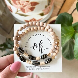 Pode incluir: Um conjunto de três pulseiras com contas de madeira natural, contas listradas em preto e dourado e pingentes de coração dourados. As pulseiras estão empilhadas em um cartão branco com o texto "The Oil Haven Boutique" e "oh" em fonte preta e dourada. O cartão também inclui o endereço do site "theoilhavenboutique.etsy.com" e o texto "Feito à mão em Appleton, Wisconsin."
