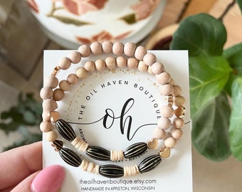Può includere: Un set di tre braccialetti con perline in legno naturale, perline a strisce nere e dorate e ciondoli a forma di cuore dorati. I braccialetti sono impilati su una carta bianca con il testo "The Oil Haven Boutique" e "oh" in caratteri neri e dorati. La carta include anche l'indirizzo del sito web "theoilhavenboutique.etsy.com" e il testo "Fatto a mano ad Appleton, Wisconsin."