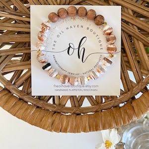 May include: A brown and white agate stone bracelet with gold accents. The bracelet is made with wooden beads and is displayed on a white card with the text "THE OIL HAVEN BOUTIQUE oh AROMATHERAPY JEWELRY NATURAL PRODUCTS theoilhavenboutique.etsy.com HANDMADE IN APPLETON, WISCONSIN".