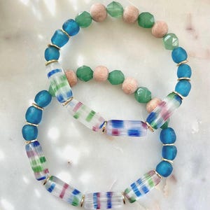 May include: Two bracelets with blue, green, and white glass beads. The top bracelet has wooden beads and the bottom bracelet has clear glass beads with stripes.