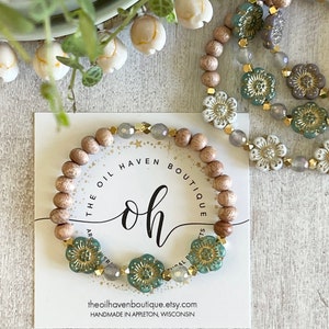 Pode incluir: Um bracelete de contas de madeira com detalhes florais verdes e dourados. O bracelete está sobre um cartão branco com o texto "The Oil Haven Boutique" e "oh" em uma fonte estilizada.