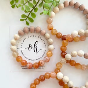 Op de afbeelding: Twee armbanden gemaakt van natuurlijke stenen en houten kralen. De armbanden liggen op elkaar. De bovenste armband is gemaakt van lichtbruine houten kralen en de onderste armband is gemaakt van oranje en witte agaat kralen. De armbanden liggen op een witte achtergrond met een visitekaartje waarop "The Oil Haven Boutique" staat, met de tekst "Aromatherapy Jewelry - Natural Products".