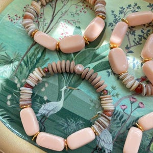 May include: Three stretch bracelets featuring smooth, rectangular light pink beads, small tan wooden disc beads, and iridescent white pearls. Each bracelet is accented with small gold-toned metal spacers between the beads, creating a uniform pattern of alternating shapes and textures throughout the circular designs.