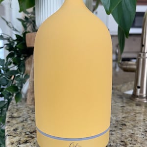 Puede incluir: Un difusor de aceites esenciales amarillo con una parte superior redondeada y una base gris claro. El difusor tiene el texto "Oil the Haven" en una fuente cursiva en la base. El difusor est&aacute; sobre una encimera moteada con plantas verdes en el fondo.