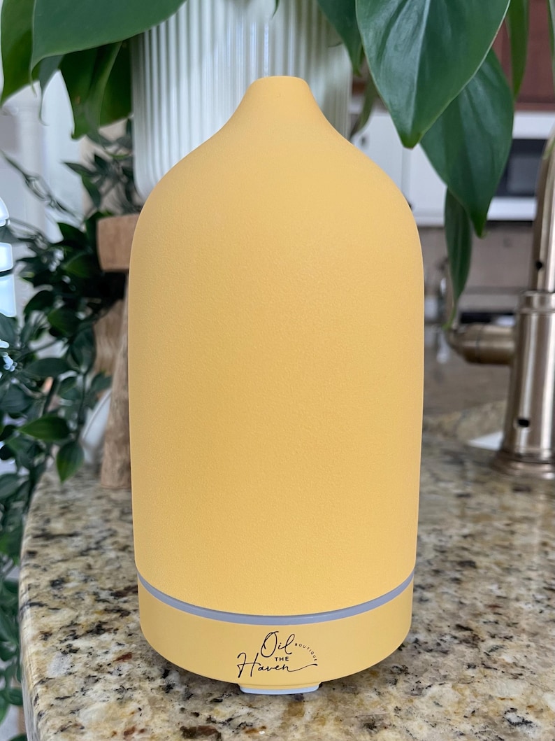 Puede incluir: Un difusor de aceites esenciales amarillo con una parte superior redondeada y una base gris claro. El difusor tiene el texto "Oil the Haven" en una fuente cursiva en la base. El difusor est&aacute; sobre una encimera moteada con plantas verdes en el fondo.