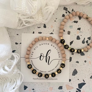May include: Two wooden bead bracelets with gold accents. One bracelet spells "BREATHE" in black letters and the other spells "LOVE" in black letters. The bracelets are on a white card with the text "The Oil Haven Boutique" and "Aromatherapy Jewellery - Natural Products".
