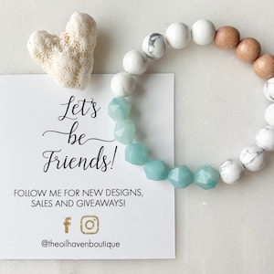May include: A beaded bracelet with white, light brown, and light blue beads. The bracelet is next to a white card with the text "Let's be Friends!" and social media handles. A white coral-shaped object is also present.