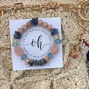 May include: A beaded bracelet featuring a variety of natural stones and wooden beads. The bracelet includes pink, blue, and gray stones, with gold-colored square accents. The bracelet is displayed on a white card with the text "The Oil Haven Boutique".