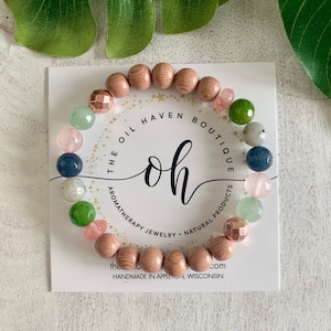 Puede incluir: Un brazalete de cuentas de madera con varias cuentas de piedras preciosas de colores, incluyendo rosa, verde, azul y gris. El brazalete está sobre una tarjeta blanca con el texto "THE OIL HAVEN BOUTIQUE oh AROMATHERAPY JEWELRY NATURAL PRODUCTS the com HANDMADE IN APPLETON WISCONSIN".
