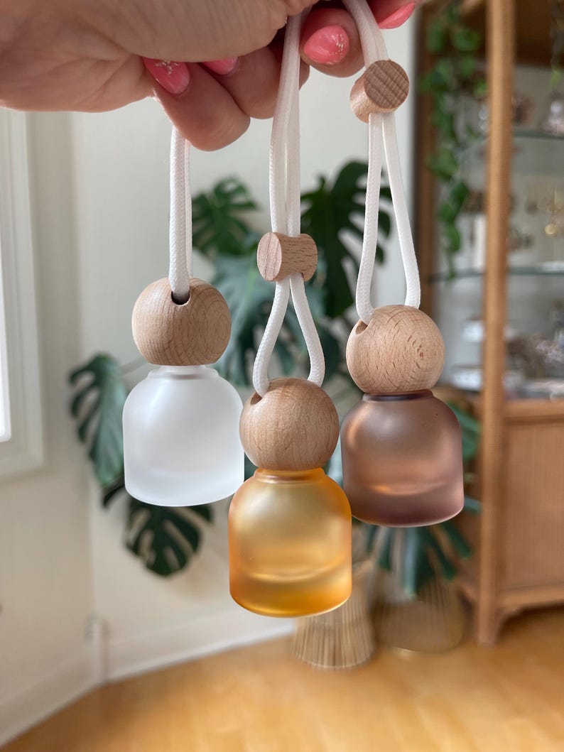 May include: Three small, round, frosted glass bottles hanging from white cords. Each bottle features a natural light-colored wooden bead cap and a matching wooden slider on the cord. The bottles are clear frosted, amber, and dark brown in color.