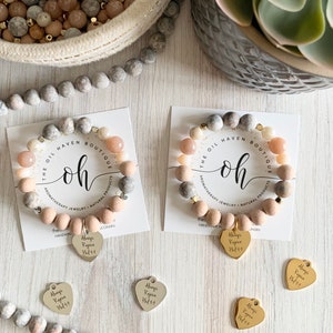May include: Two bracelets with pink and grey beads and gold accents. Each bracelet has a heart charm with the text "Always Regina" and a tag with the text "The Oil Haven Boutique OH Aromatherapy Jewellery Natural Products".