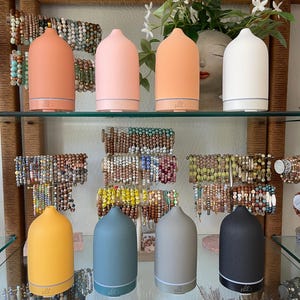 Puede incluir: Exhibici&oacute;n de ocho difusores de aceites esenciales de cer&aacute;mica en varios colores, incluyendo melocot&oacute;n, rosa, naranja, blanco, amarillo, azul, gris y negro. Los difusores est&aacute;n colocados en estantes de vidrio, con pulseras de cuentas en el fondo.