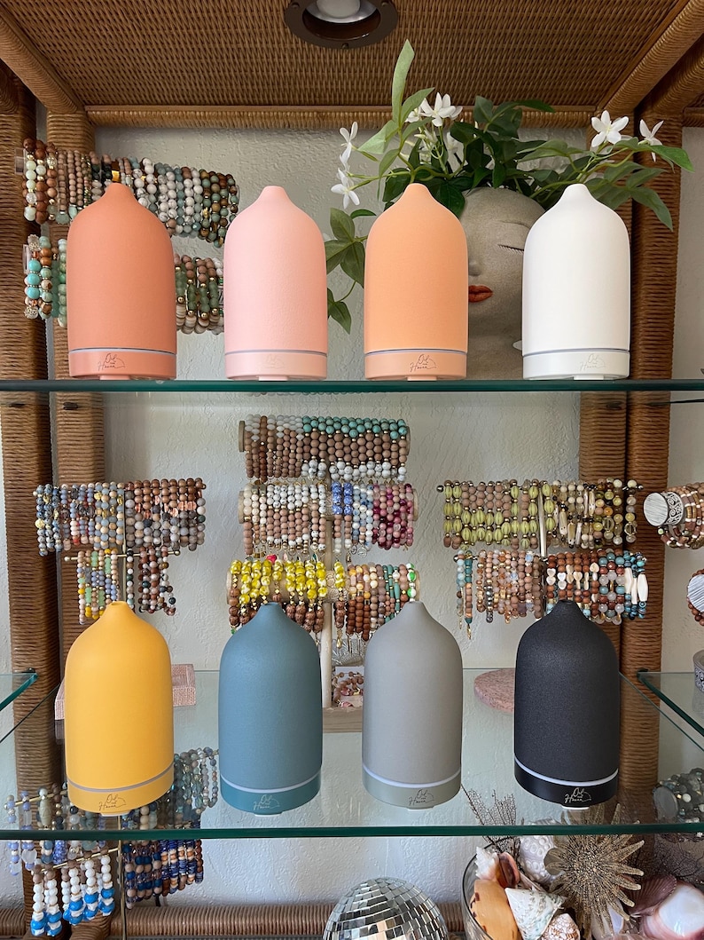 Puede incluir: Exhibici&oacute;n de ocho difusores de aceites esenciales de cer&aacute;mica en varios colores, incluyendo melocot&oacute;n, rosa, naranja, blanco, amarillo, azul, gris y negro. Los difusores est&aacute;n colocados en estantes de vidrio, con pulseras de cuentas en el fondo.