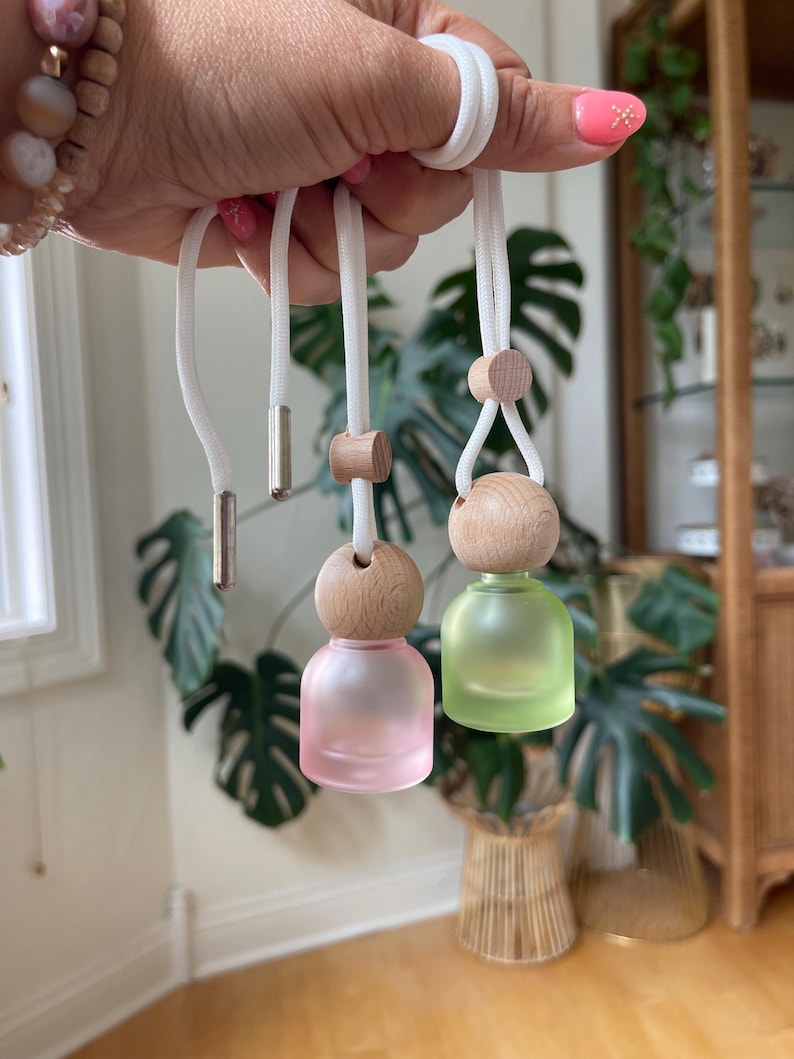 May include: Two small, round, frosted glass bottles hanging from white cords. One bottle is light pink and the other is light green. Each bottle features a natural light-colored wooden cap and a small wooden bead on the cord for adjustment.