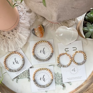 Puede incluir: Tres pulseras de cuentas de madera con detalles dorados y un diseño a rayas blanco y gris. Cada pulsera tiene una tarjeta con el texto "The Oil Haven Boutique" y "oh" en letra cursiva. Las tarjetas también incluyen el texto "theoilhavenboutique.etsy.com" y "Hecho a mano en Appleton, Wisconsin".