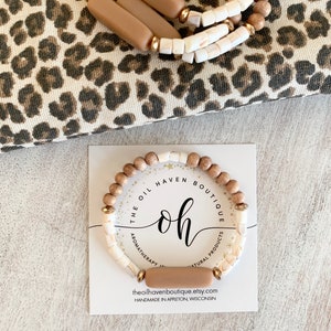 Pode incluir: Um bracelete de madeira com contas marrons e brancas, uma barra marrom com o texto "the oil haven boutique" e o texto "oh" em um círculo com as palavras "aromaterapia" e "produtos naturais".