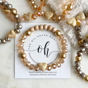 Pode incluir: Um conjunto de três pulseiras de contas douradas e marrons com pingentes de coração dourados. As pulseiras estão sobre um cartão branco com o texto "The Oil Haven Boutique, Aromatherapy Jewelry, Natural Products, theoilhavenboutique.etsy.com, Handmade in Appleton, Wisconsin".
