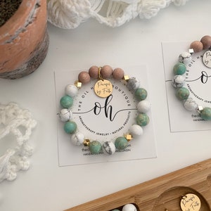 Può includere: Un braccialetto in pietra turchese e bianca con dettagli dorati e un ciondolo dorato che dice "Pureful by Faith". Il braccialetto è su una carta bianca con le parole "The Oil Boutique" e "Aromatherapy Jewelry - Natural Products" stampate su di essa.