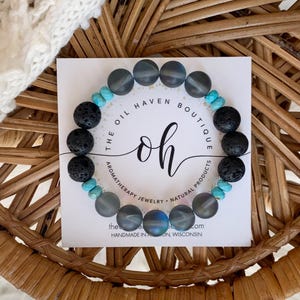 May include: A beaded bracelet featuring gray, black, and turquoise stones. The bracelet is displayed on a white card with the text "THE OIL HAVEN BOUTIQUE" and "oh" in a cursive font. The bracelet is on a woven wicker surface.