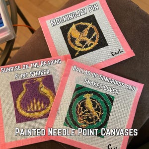 May include: Three square needlepoint canvases with pink taped edges. The designs feature a gold Mockingjay pin on black, a gold flint striker on purple, and a gold songbird on a green and black background. Text labels include Mockingjay Pin, Sunrise on the Reaping Flint Striker, Ballad of Songbirds and Snakes Cover, and Painted Needle Point Canvases.