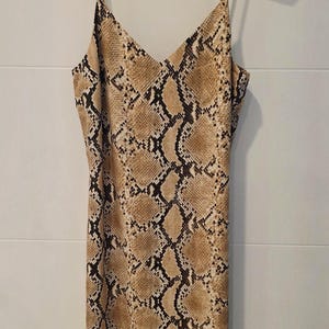 Könnte beinhalten: Midikleid mit Spaghettiträgern und Schlangenmuster in Braun- und Schwarztönen. Das Kleid hat einen V-Ausschnitt. Es hängt an einem Kleiderbügel mit Leopardenmuster.