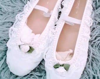 May include: A pair of white satin ballet flats with lace trim and pearl accents. The flats have a green bow on the toe and a white satin rose on the side.