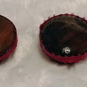 May include: Two round, decorative buttons. One button has a brown and black striped pattern, the other has a dark brown and gold pattern. Both buttons have a red border with a stitched edge.