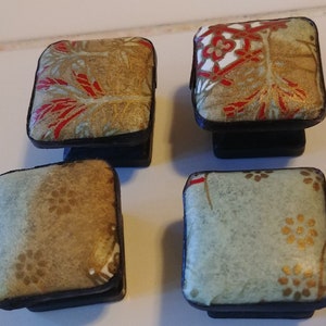 May include: Four square cabinet knobs with decorative fabric tops. The fabric patterns include gold, red, and white floral designs on a tan background. The knobs have black bases and are set against a white tiled surface.