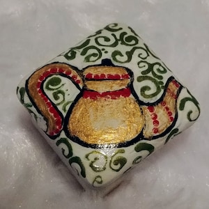 May include: A hand-painted, square-shaped decorative stone featuring a gold teapot design. The teapot has red and gold accents, set against a white background with green swirl patterns. The stone is a unique piece of art.