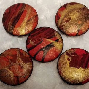 May include: Five round, decorative buttons with a marbled design. The buttons feature a mix of red, gold, and black colors. The buttons are arranged on a white, textured surface.