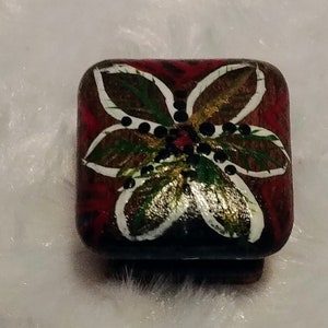 May include: A square, wooden cabinet knob with a hand-painted floral design. The flower has white petals with gold accents, green leaves, and black dots, set against a dark red background. The knob is likely for furniture or decorative use.