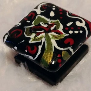 May include: A square, black decorative object with a floral design. The design features red, white, and green accents on a black background. The object is sitting on a smaller, black square base.