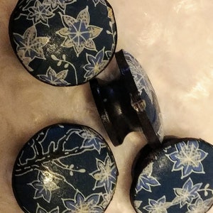 May include: A collection of dark blue, round wooden drawer pulls with a floral design in white and light blue. The pulls have a slightly domed shape and are arranged on a soft, light-colored surface.