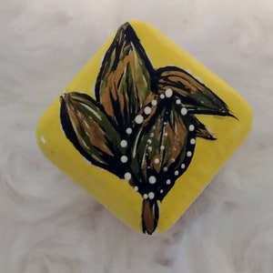 May include: A hand-painted, yellow, diamond-shaped rock featuring a botanical design. The design includes green and brown leaves with white dot accents. The rock is a unique piece of art.