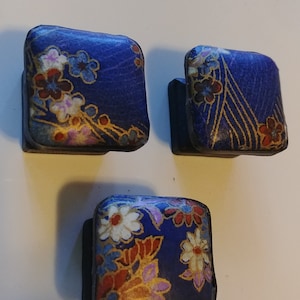 May include: Three square cabinet knobs with a dark blue background and floral patterns in white, red, and gold. The knobs are arranged in a triangular pattern, showcasing their decorative design.