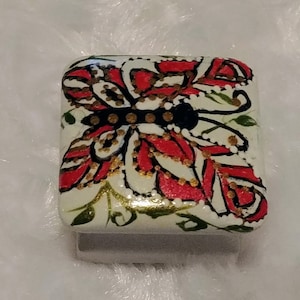 May include: Square, hand-painted decorative object with a butterfly design. The butterfly has red and white wings with gold accents and a black body. The background is off-white with green leaf accents.