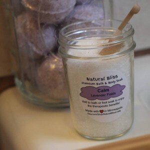 Peut inclure: Un pot en verre transparent rempli de sels de bain blancs et une petite cuillère en bois. Le pot porte une étiquette indiquant "Natural Bliss Calm Lavender Fields". Un pot plus grand avec des bombes de bain violettes est en arrière-plan.