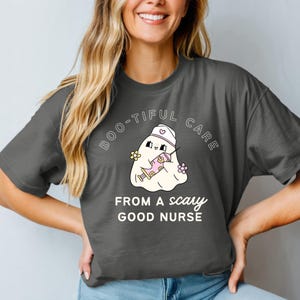 Peut inclure: T-shirt gris foncé avec le texte "BOO-TIFUL CARE FROM A SCARY GOOD NURSE". Le t-shirt présente un fantôme de dessin animé portant un chapeau d'infirmière et tenant une seringue. Le fantôme est entouré de fleurs.