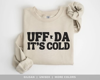 May include: A beige sweatshirt with the phrase "UFF DA IT'S COLD" in bold black letters. The design includes a silhouette of the state of Minnesota. The sleeves are rolled up, and the sweatshirt is laid flat.