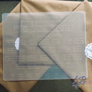 Puede incluir: Una hoja de práctica de borrado en seco de plástico transparente con el texto "Hello, my name is Ela Mae Reed and I can write..." en la parte superior. La hoja presenta una cuadrícula de letras minúsculas y números para trazar.