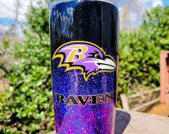 May include: A black and purple glitter tumbler with the Baltimore Ravens logo. The logo features a purple raven with a gold "B" on its chest and the words "Baltimore Ravens" in gold lettering.