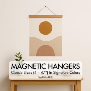 May include: A beige and brown abstract art print with a mustard yellow sun and a brown semi-circle, displayed with a wooden magnetic hanger. The print is hanging on a white wall. The text "MAGNETIC HANGERS" is displayed below the print.